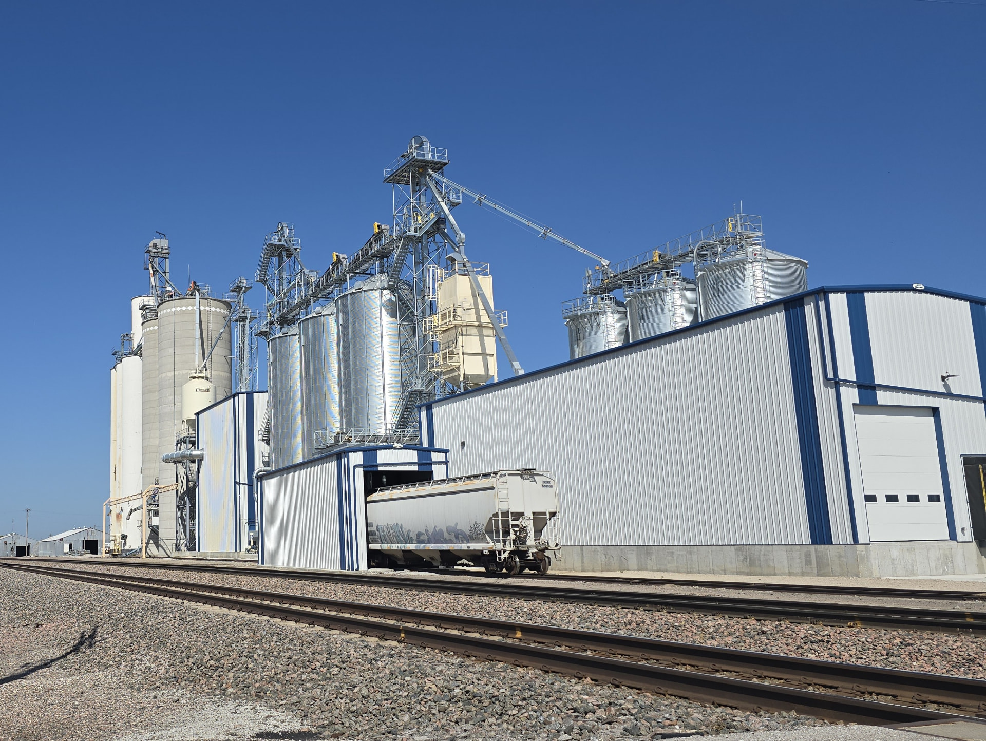 Large grain elevator and warehouse complex with white corrugated siding alongside multiple railroad tracks